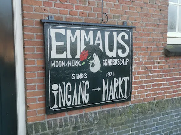 Vintage uithangbord aan bakstenen gevel met witte letters en rood logo, 'EMMAUS' met 'SINDS 1971 INGANG MARKT'