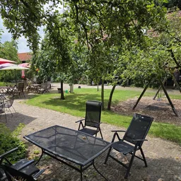 Tuinterras met zwarte loungestoelen en tafel onder hangende wilgen, rode parasol op achtergrond