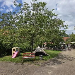 Speeltuin met glijbaan en zandkuil onder grote bomen op terrein van kringloopwinkel