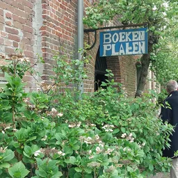 Ingang van kringloopwinkel Boeken Plaats met blauwe uithangbord, omgeven door groen en bloeiende planten