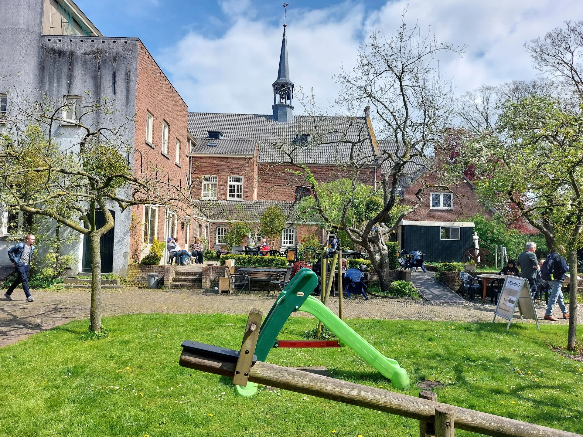 Binnentuin met gras, speeltoestellen en oude fruitbomen, omgeven door bakstenen gebouwen