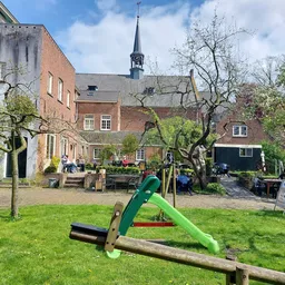 Binnentuin met gras, speeltoestellen en oude fruitbomen, omgeven door bakstenen gebouwen