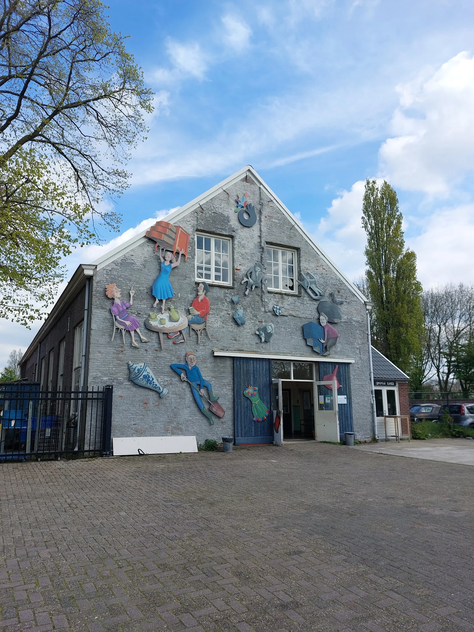 Grijstenen gevel van kringloopwinkel Emmaus Langeweg met kleurrijke muurschildering van spelende kinderen en blauwe deuren