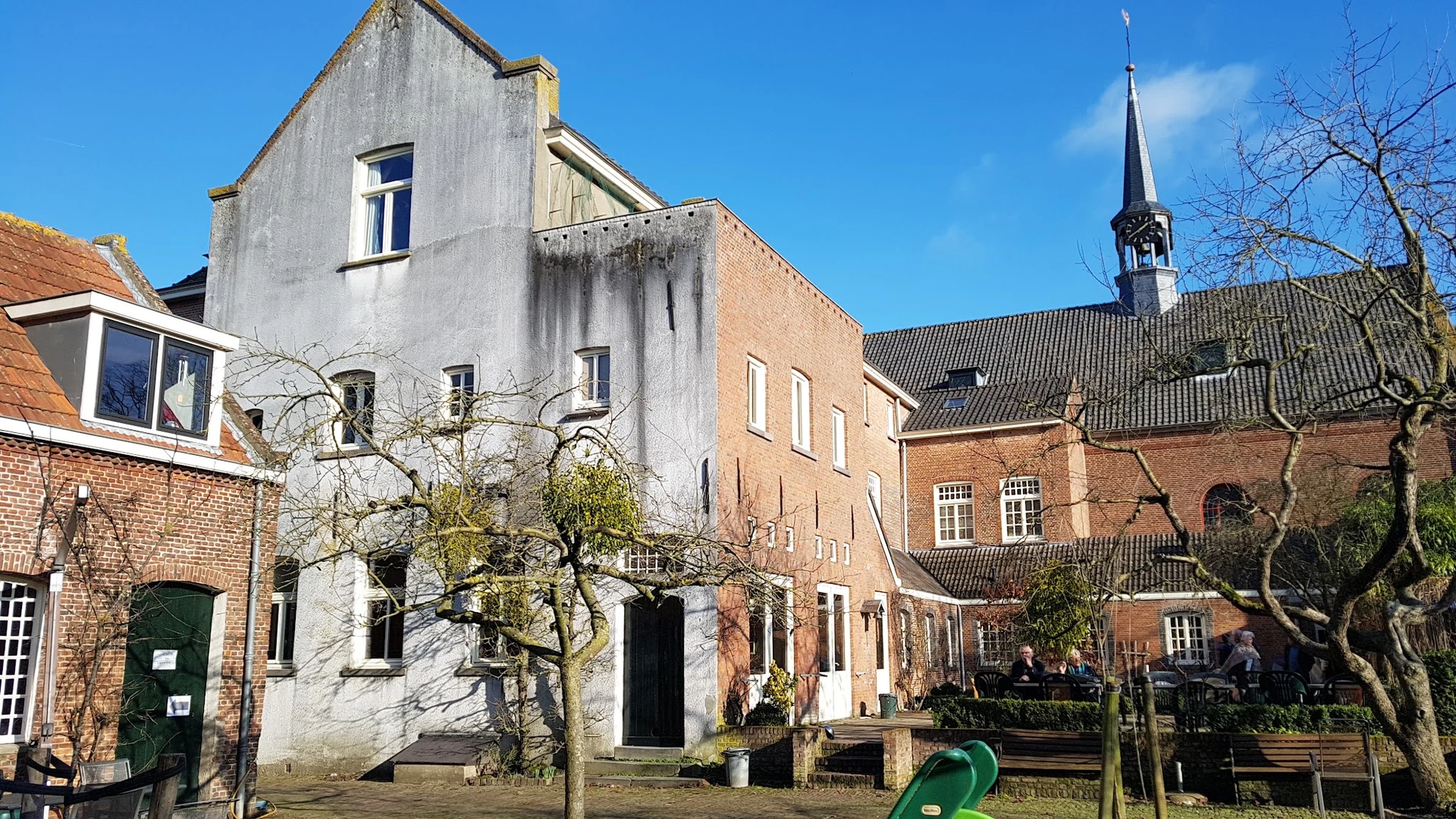 Voorkant van Emmaus kringloopwinkel met witgeschilderd en bakstenen gebouw, toegangsdeur en ramen, kerk op achtergrond