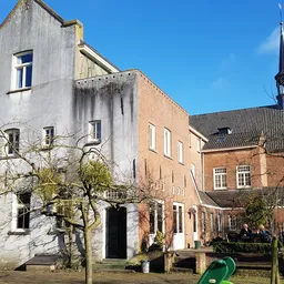 Voorkant van Emmaus kringloopwinkel met witgeschilderd en bakstenen gebouw, toegangsdeur en ramen, kerk op achtergrond