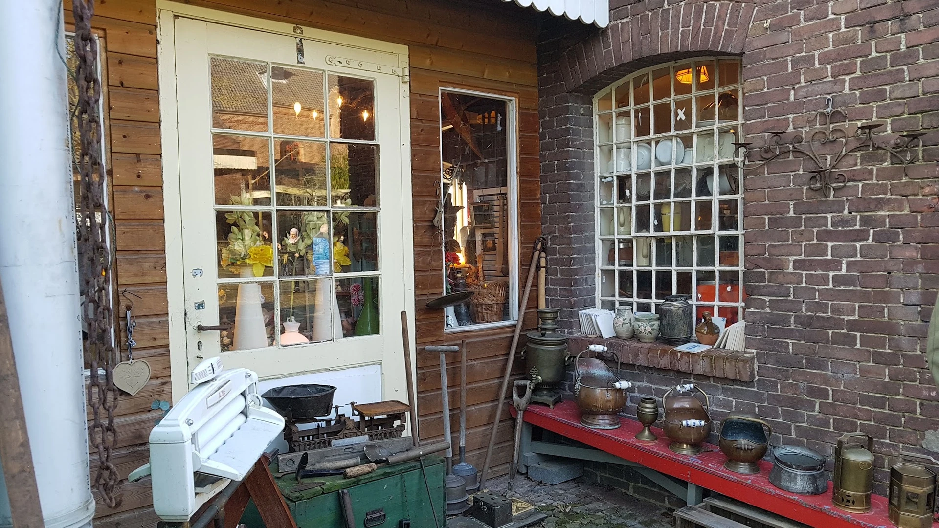 Uitgestalde huishoudelijke artikelen en keramiek op rode en groene werkbanken buiten de winkel