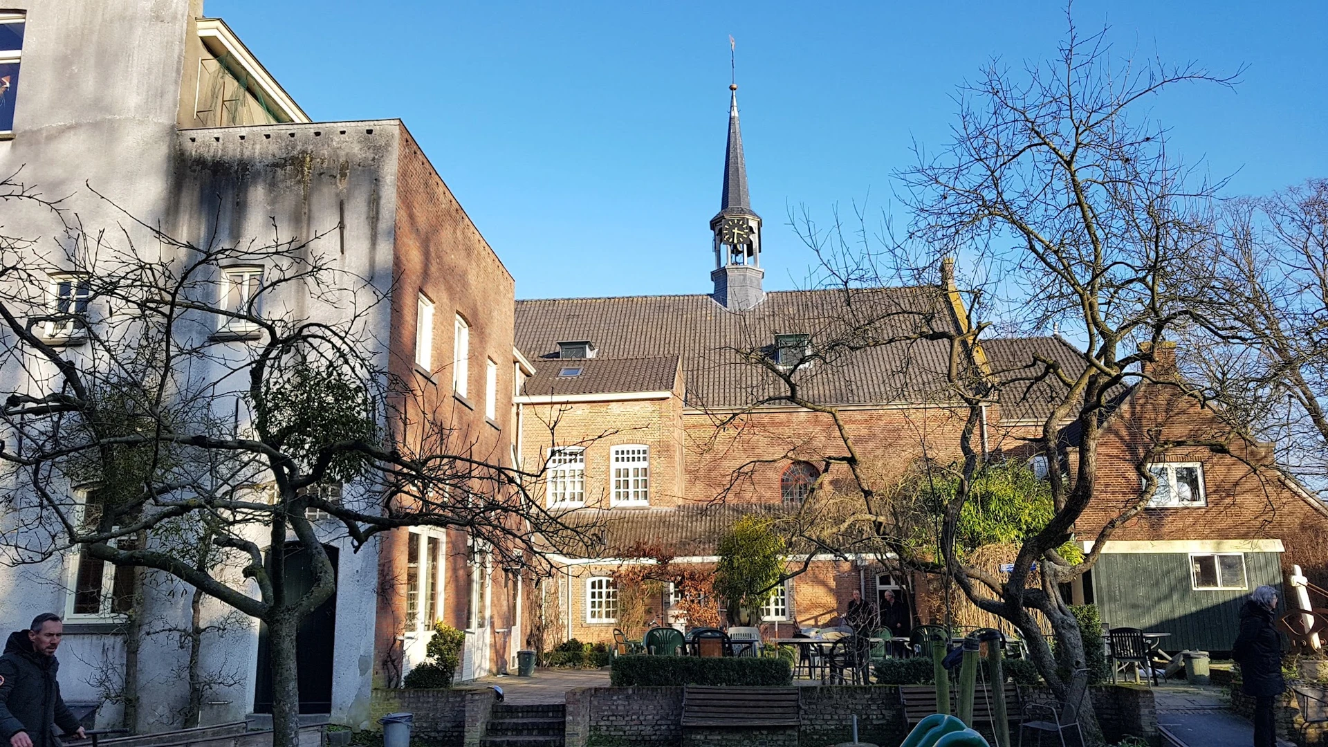 Voorgevel van een bakstenen kerkgebouw met spits en daarnaast een gele bakstenen vleugel, omgeven door kale bomen