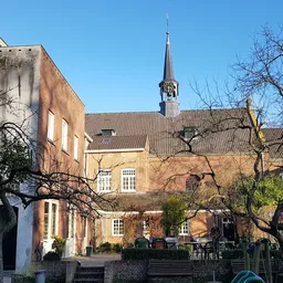 Voorgevel van een bakstenen kerkgebouw met spits en daarnaast een gele bakstenen vleugel, omgeven door kale bomen