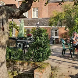 Terras met tafels en stoelen onder een oude boom bij een bakstenen gebouw