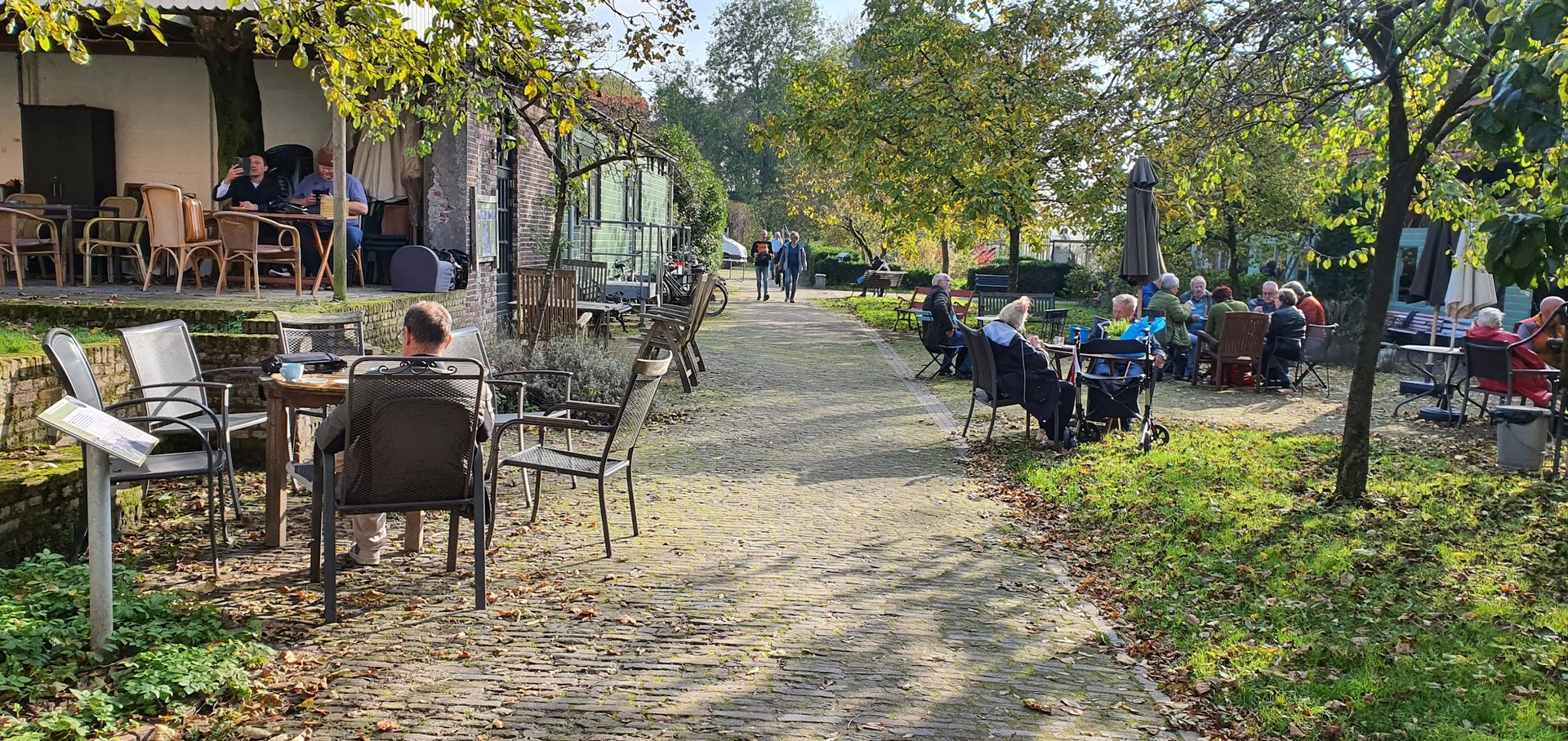 Bezoekers zitten aan tafels op een terras onder grote bomen in een groen park