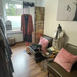 Winkelinterieur met kledingrekken, zithoek met roze kussens, kleding op hangers en uitzicht op raam