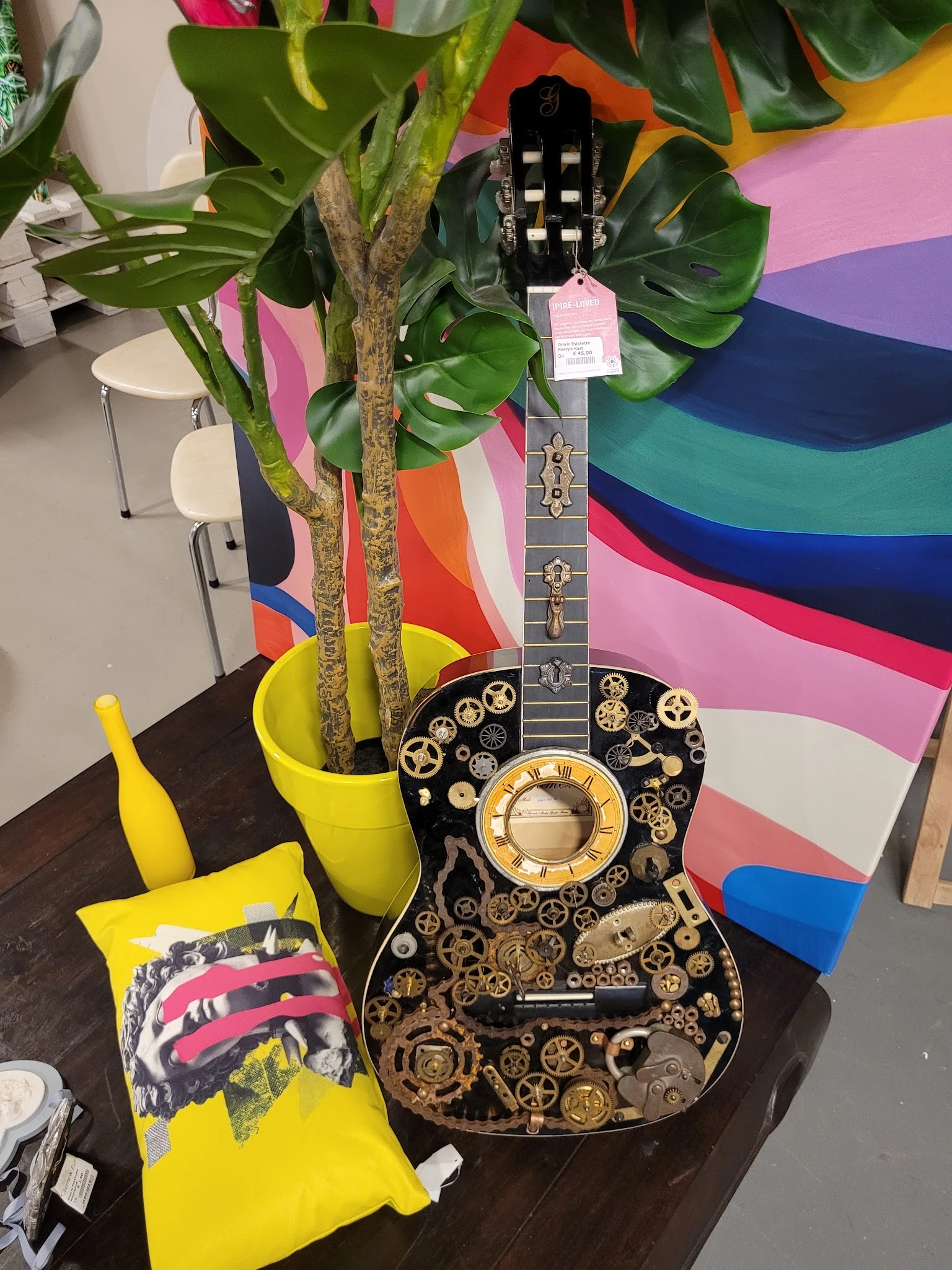 Decoratieve gitaar met zwart lichaam, gouden tandwielen en klokwerk motief, naast gele plantenpotten en plant.