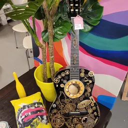 Decoratieve gitaar met zwart lichaam, gouden tandwielen en klokwerk motief, naast gele plantenpotten en plant.