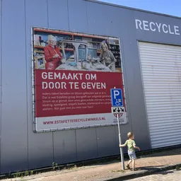 Groot reclamebord aan gevel met afbeelding en Nederlandse tekst, gedeeltelijk zichtbare winkelwand