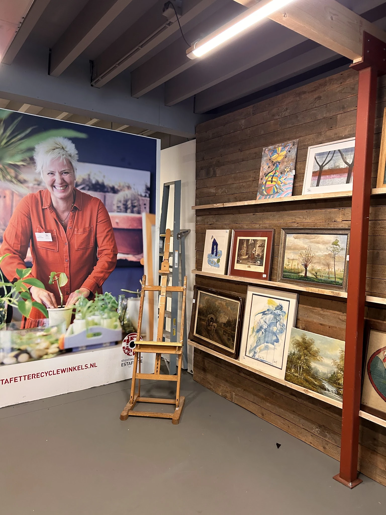Winkelinterieur met houten wandrekken vol kunstwerken en schilderijen, naast een groot portretposter van een vrouw in oranje 