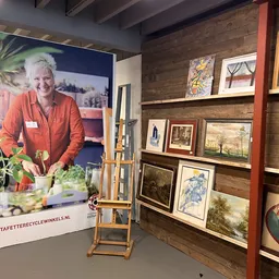 Winkelinterieur met houten wandrekken vol kunstwerken en schilderijen, naast een groot portretposter van een vrouw in oranje 