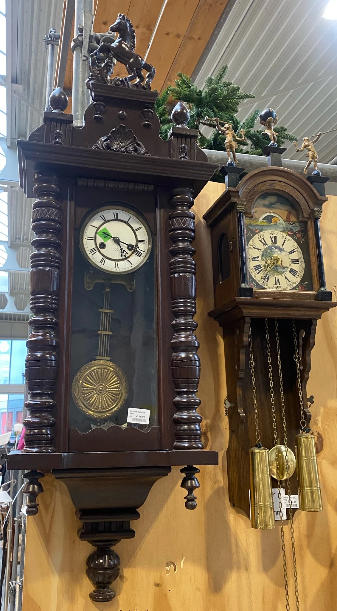 Twee houten wandklokken met Romeinse cijfers en slingers, tentoongesteld in de kringloopwinkel
