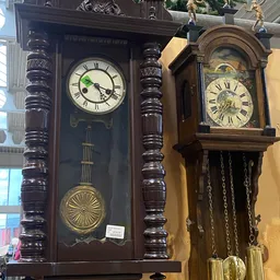 Twee houten wandklokken met Romeinse cijfers en slingers, tentoongesteld in de kringloopwinkel