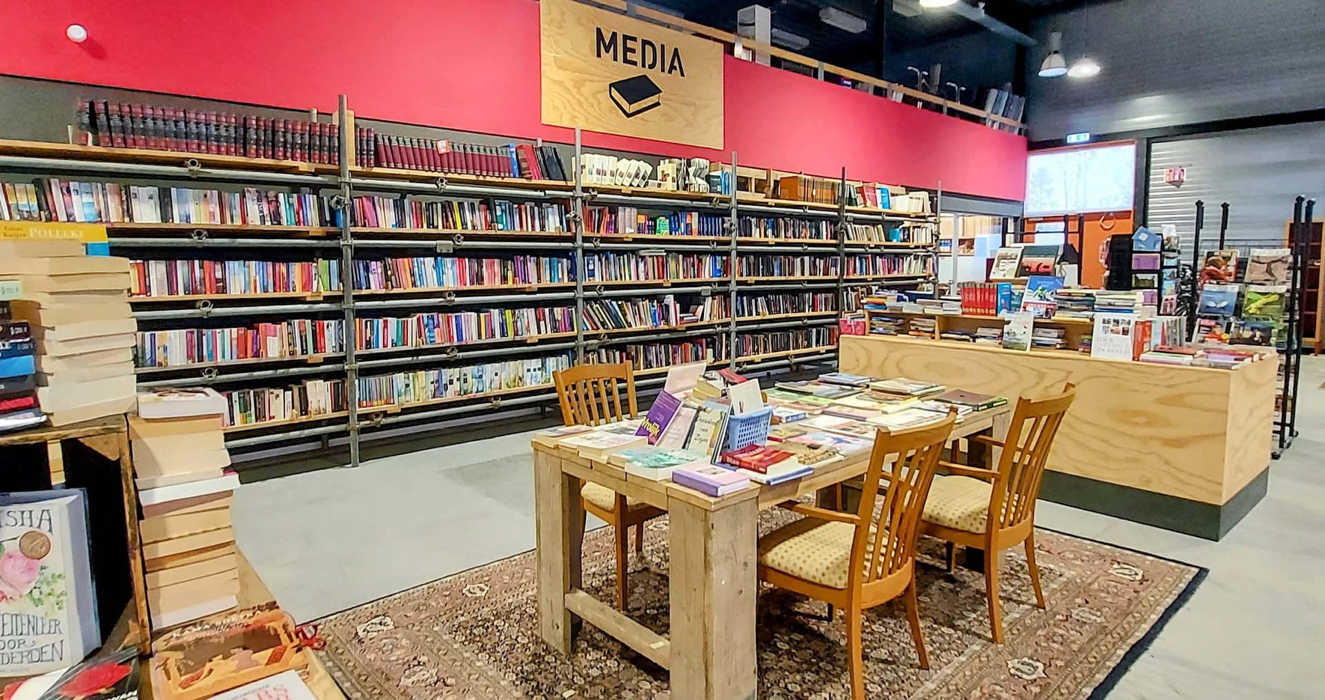 Mediaafdeling in kringloopwinkel met boekenrekken tegen roze wand en houten tafel met stoelen waarop boeken uitgestald