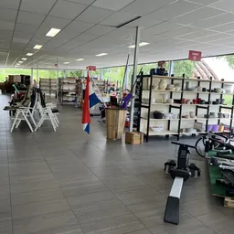 Ruim winkelbinnenruimte met stellingen vol huishoudelijke artikelen, enkele klanten, witte stoelen en sportapparatuur op de v