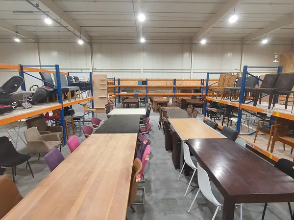 Overzicht van winkelruimte met houten en donkere eettafels, stoelen in verschillende kleuren, en stellingen met huisraad op d