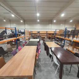 Overzicht van winkelruimte met houten en donkere eettafels, stoelen in verschillende kleuren, en stellingen met huisraad op d