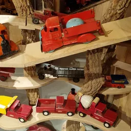 Speelgoedvoertuigen in verschillende kleuren uitgestald op houten planken rond boomstammen