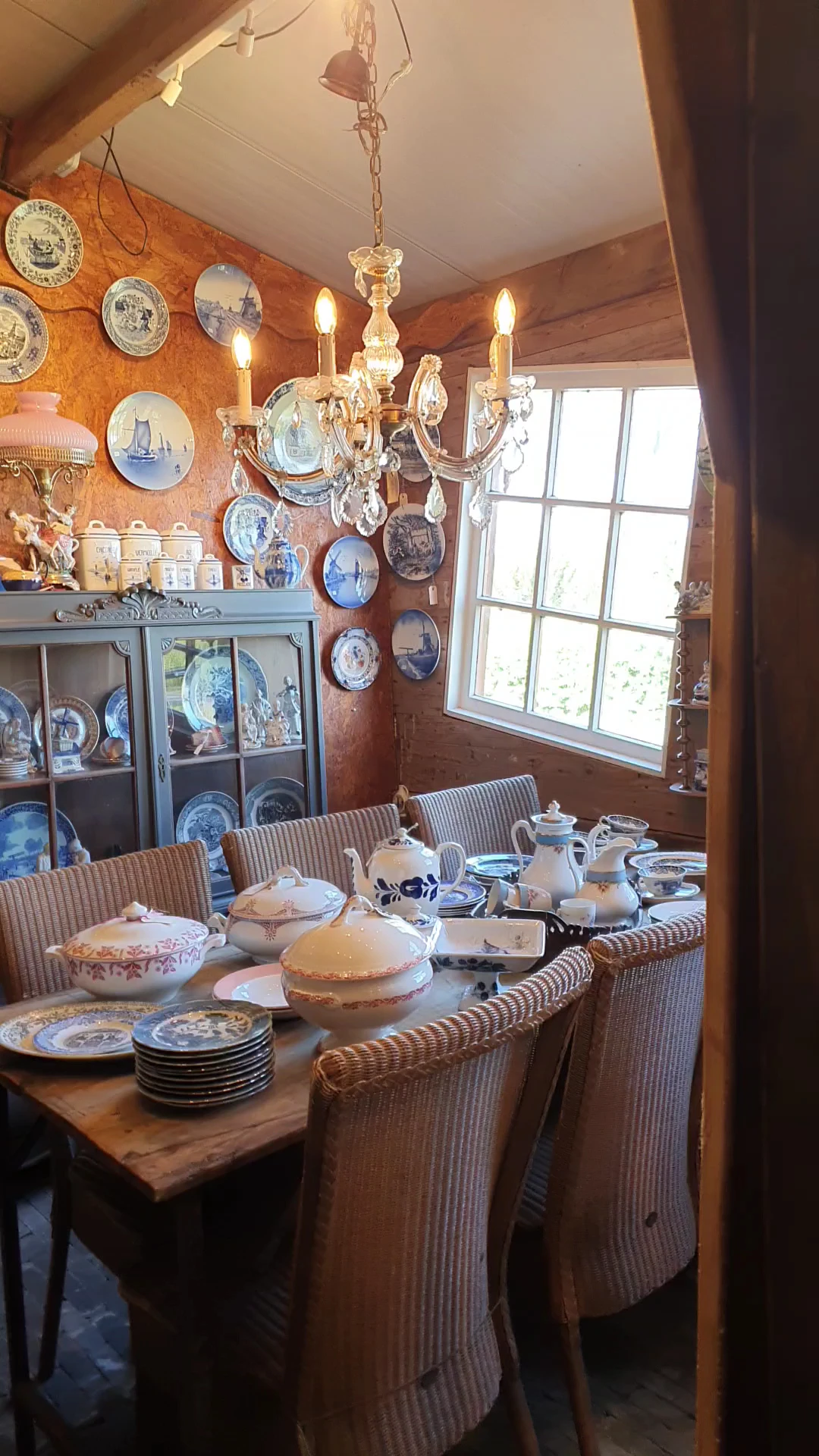 Eetkamer met gedeckte tafel, blauw-witte borden aan de muur, glazen vitrinekast en raam met uitzicht