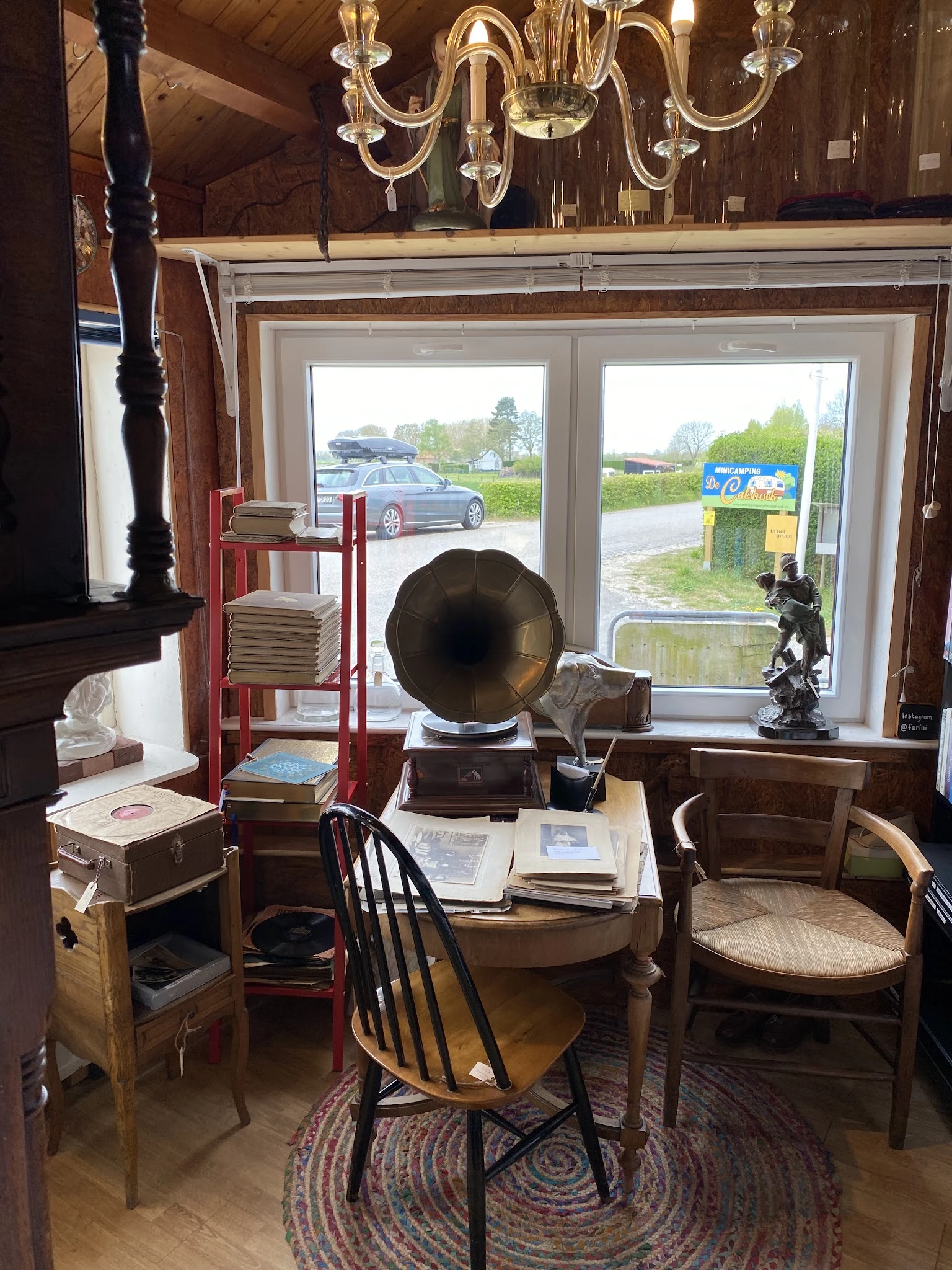 Winkelinterieur met vintage meubels, grammofoon, boekenrekken en grote ramen met uitzicht op parkeerplaats