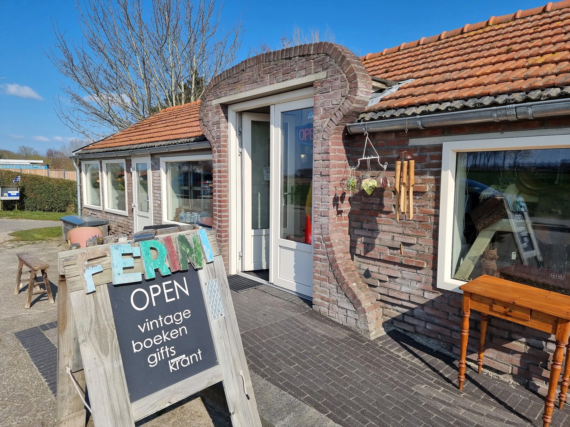 Bakstenen gevel van vintage- en boekhandel met terracotta dakpannen, grote ramen en open deur, met OPEN-bord op het terras