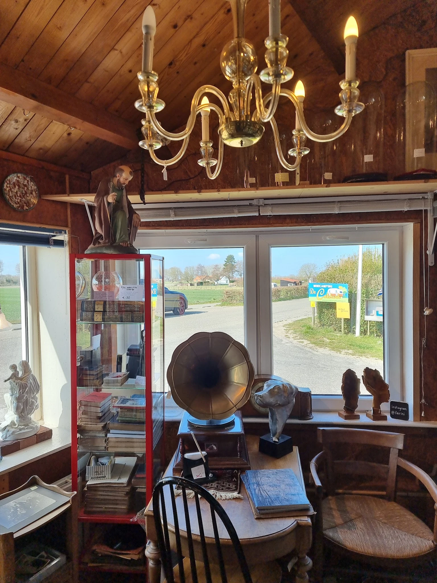 Winkelinterieur met vintage decorstukken, gramofoon, beelden en religieuze voorwerpen langs raam met zicht op landweg