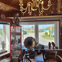 Winkelinterieur met vintage decorstukken, gramofoon, beelden en religieuze voorwerpen langs raam met zicht op landweg