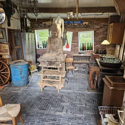 Winkelbinnenkant met boeddhabeeldhouwwerk, vintage meubels, blauwe decoratieve vat en diverse tweedehands artikelen op herrin