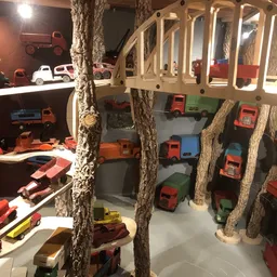 Winkelbinnenruimte met speelgoedtreinen en voertuigen uitgestald tussen boomstammen, met een houten loopbrug erboven