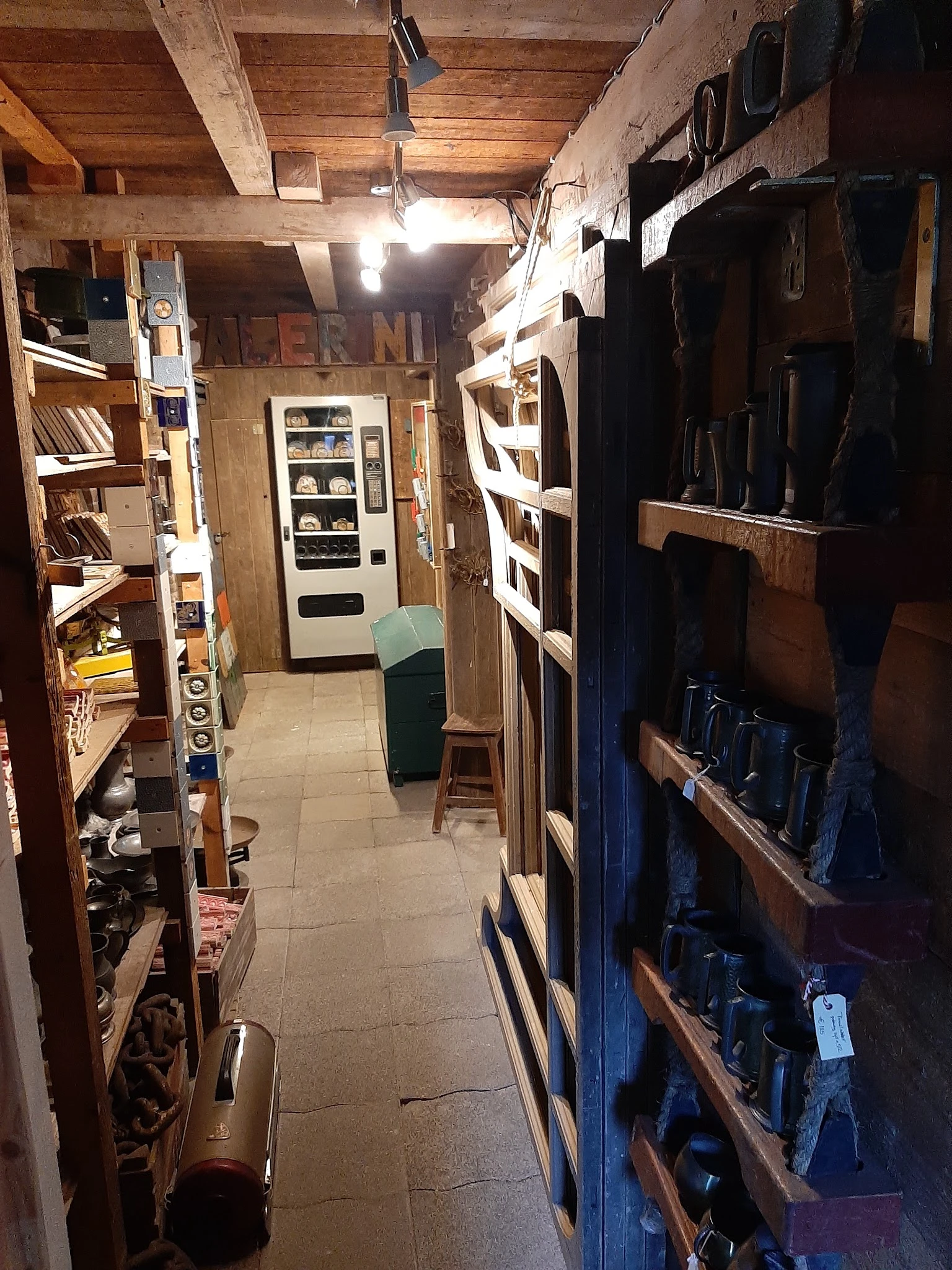 Nauwe winkelgang met boekenkast links, automaat en groen zitje in het midden, en stellingen met artikelen rechts