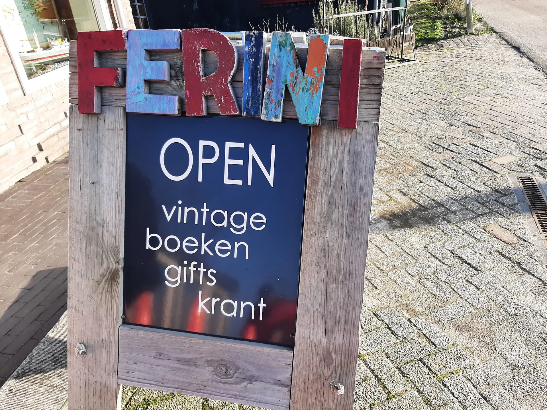 Houten bord met gekleurde letters 'FERINI' en zwart informatiebord met 'OPEN' en winkelomschrijving op straathoek