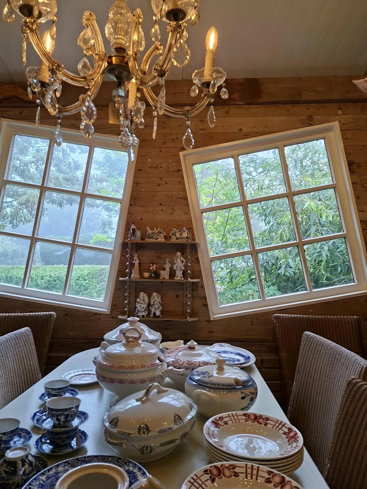 Houten kamer met twee grote raampartijen, serviezen op tafel, decoratieve beeldjes op houten planken aan muur