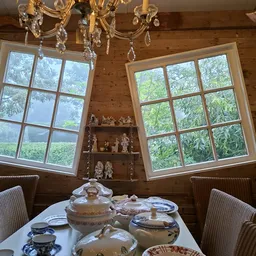 Houten kamer met twee grote raampartijen, serviezen op tafel, decoratieve beeldjes op houten planken aan muur