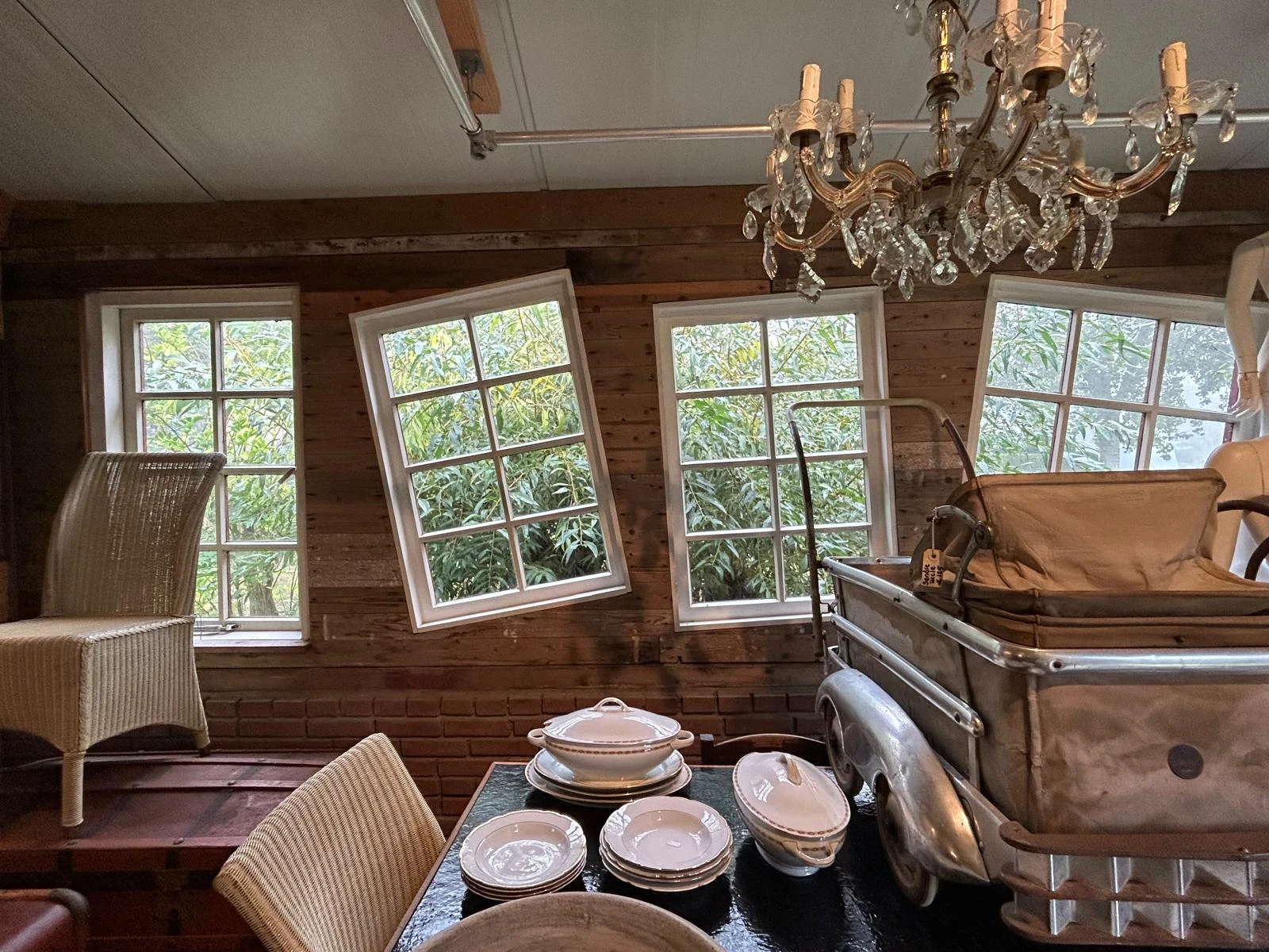 Woonkamer met houten wanden, grote ramen, kroonluchter, vintage meubels en serviesgoed op tafel