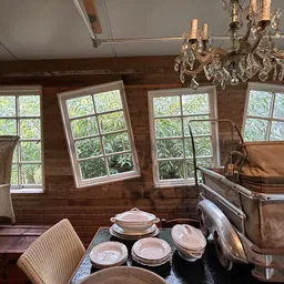 Woonkamer met houten wanden, grote ramen, kroonluchter, vintage meubels en serviesgoed op tafel