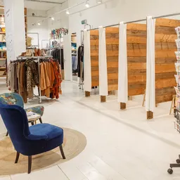 Winkelvloer met kledingrekken, houten scheidingswanden, blauwe stoel op rond tapijt en displaykasten met merchandise