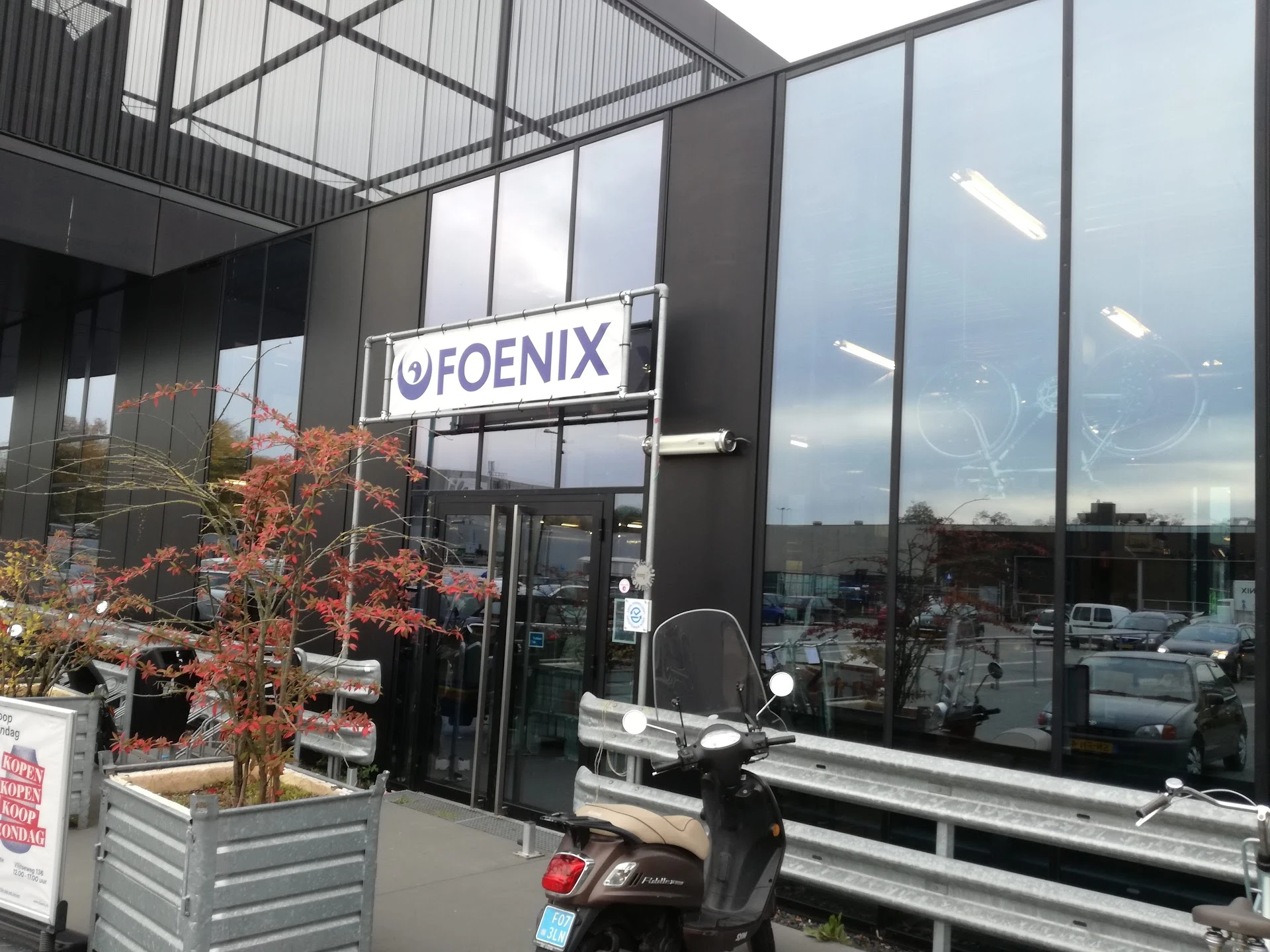 Glazen gevel van kringloopwinkel met Foenix-logo, plantenbakken en parkeerplaats zichtbaar