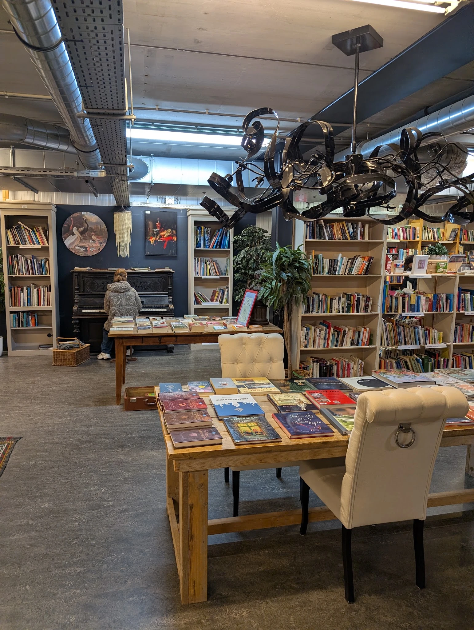 Ruim interieur van kringloopwinkel met boekenkasten, piano, houten tafels met uitgelegde boeken en gele stoelen