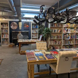 Ruim interieur van kringloopwinkel met boekenkasten, piano, houten tafels met uitgelegde boeken en gele stoelen