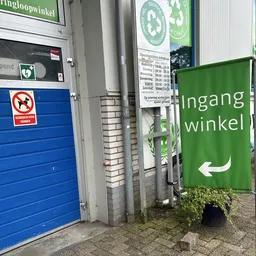 Ingang van kringloopwinkel met blauwe deur, no-dogs-bord en groen winkelnaambord met pijl