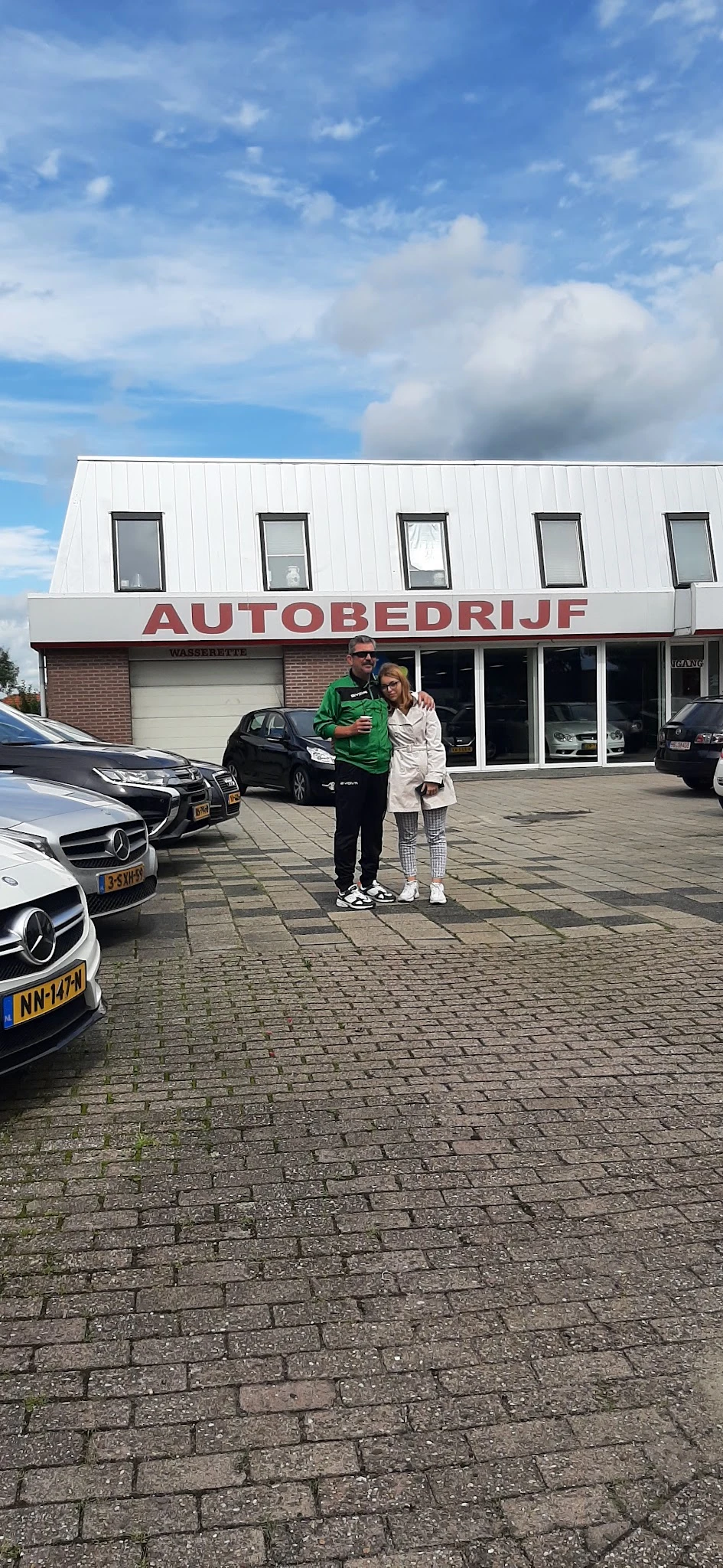 Voorgevel van autobedrijf met twee personen staand op het parkeerterrein, auto's zichtbaar