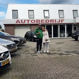 Voorgevel van autobedrijf met twee personen staand op het parkeerterrein, auto's zichtbaar