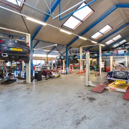 Garage-werkplaats met auto op hefbrug, werkbanken en gereedschap onder helder verlichte hal met blauwe spanten