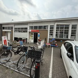 Voorkant van kringloopwinkel met parkeerplaats, fietsen en witte auto