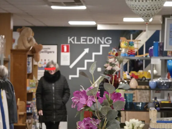 Interieur van kringloopwinkel met kleding, decoratie en huishoudspullen op stellingen en display tegen donkere wand met 'KLED